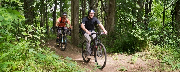 two men wearing helmets, riding mountain bikes on a forested trail