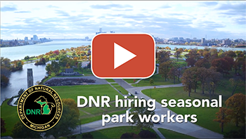 thumbnail play button image from DNR video about hiring seasonal park workers, showing a worker helping a customer at park entrance