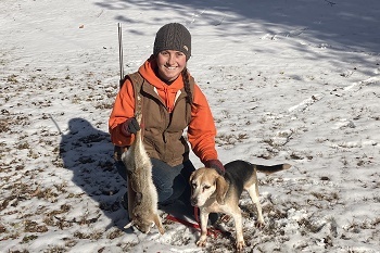 a smiling, kneeling woman, dressed in sweatshirt, vest and hat, holds a gun and a harvested rabbit, her beagle nearby in the snow