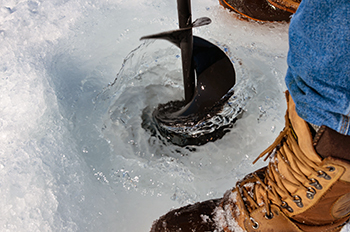 A close-up photo shows an ice angler drilling a hole through the ice on Lake Superior with an auger.