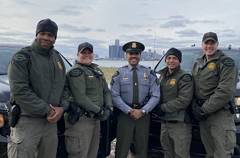 a small group of uniformed, smiling male and female Michigan DNR conservation officers, against a city backdrop