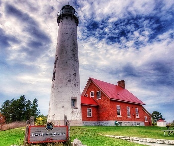 The red-orange brick and red roof of the Tawas Point Lighthouse keeper's quarters and the stately white light tower against a deep blue, cloudy sky