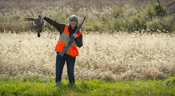 female hunter in orange vest with shotgun holding up a pheasant