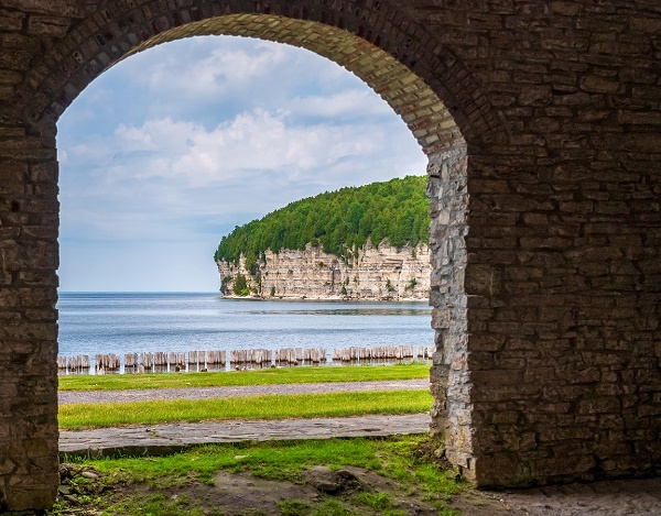 the view looking out from a covered stone archway, onto a large harbor, with a backdrop of tall, yellowed limestone cliffs overgrown with brush