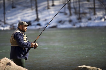a bearded man wearing a fishing vest and baseball cap holds a fishing rod over the river bank, against a snowy, treed background