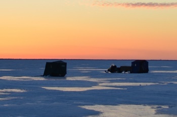 shadowed ice shanties and a snowmobile in the distance on iced-over lake, with an orange sky, few clouds, behind them