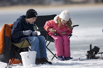 little boy in dark blue coat, little girl in pink coat and snow pants, holding fishing poles and sitting in chairs over an ice fishing hole