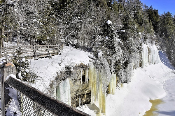 view from a wooden, railed walkway that wraps to the left, overlooking frozen waves of green-brown water, with snow-covered pines surrounding