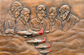 Carved, copper-colored relief image showing five uniformed pilots looking down from the clouds as four red-nosed/tailed airplanes fly in formation