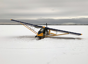 A plane with a yellow fuselage is shown sitting on the snow and ice-covered surface of Indian Lake.