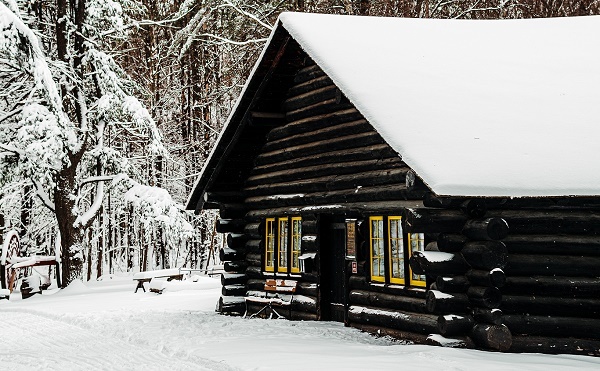 angled view of a dark brown, log cabin with yellow trim and snow-covered roof, set among snow-covered trees and a lone picnic bench out front