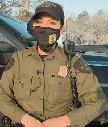 a female conservation officer wearing khaki uniform, cap and face mask, stands in front of a patrol truck, hands folded in front