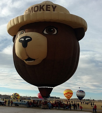 a dark brown and white hot air balloon shaped like Smokey Bear's head looms over an open, grassy area filled with other hot air balloons on ground