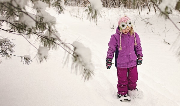 young, smiling blond girl in purple snowsuit and pink knit hat watches the ground as she snowshoes down a snow-covered, forested trail