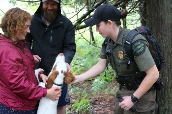 a smiling, red-haired woman, a bearded man in hoodie and a female conservation officer laugh while petting a white and brown dog jumping up 