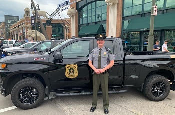 A uniformed, male conservation officer stands in front of his  black DNR patrol truck, next to Comerica Park baseball stadium in Detroit
