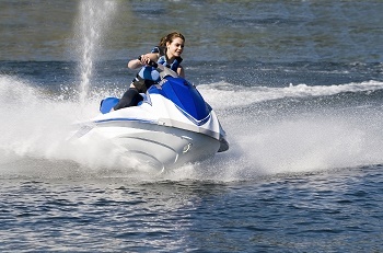 a young woman with light brown hair, wearing black and blue life vest, steers a blue and white Jet Ski making waves on dark blue water