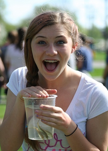 young girl with light brown braid on one side, mouth open wide in a smile, holding a see-through cup of young salmon up near her face