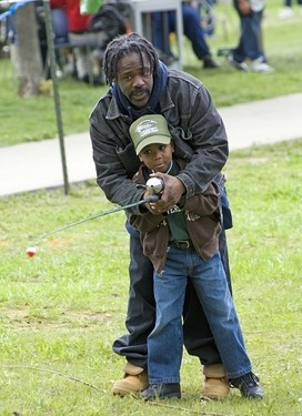 a Black man and little boy, both dressed in dark jeans and jackets, boy in a khaki baseball cap, hold a fishing pole while standing on a grassy shore