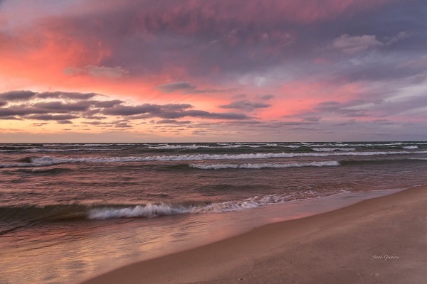 dark, gray waves with white foam roll toward brown, sandy shore, against backdrop of an orange, blue and tan sunset sky, with thin gray clouds 