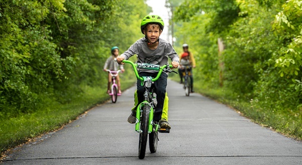young boy wearing gray T-shirt and helmet rides a neon green bike toward camera on paved, tree-lined trail, two kids farther behind him