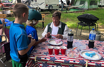 A state park campground host entertains two young children with a craft while sitting at a picnic table.
