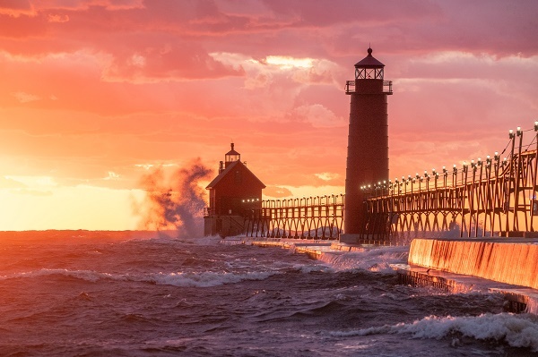 orange and pink sunset view of a concrete and steel pier connecting a tall, red light tower and a smaller lighthouse, dark gray waves crashing