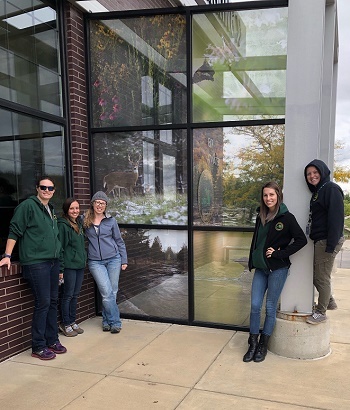 a group of smiling men and women in DNR apparel stand in front of a tall, 6x6 pane window entrance, with superimposed nature scenes on windows