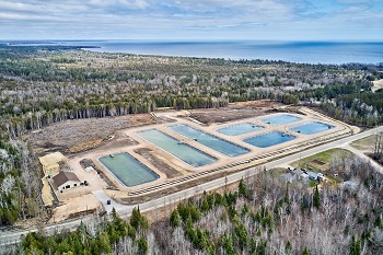 aerial view of the building, blue-lined holding pools and campus of Thompson State Fish Hatchery, surrounded by forest, Lake Michigan in background