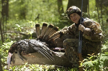 A young male hunter dressed in full camouflage and holding a shotgun upright, crouches down next to a large turkey, in the full woods
