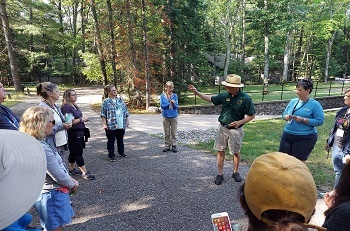 A group of men and women dressed in shorts, T-shirts, khakis and other outdoor gear, listen as a man in a green DNR shirt and brimmed hat speaks.