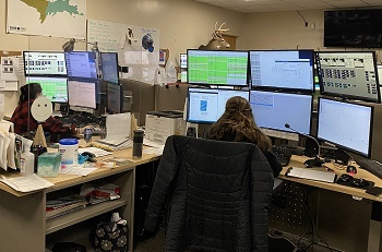 back view of two women, seated at desks with stacked computer screens, microphones and other office supplies