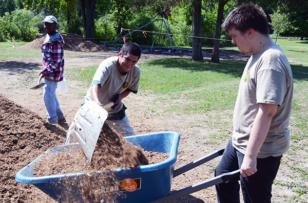 Three youths work on an outdoor project, one with a shovel, another with a wheelbarrow and a third walking past.