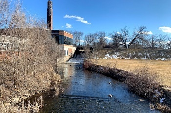 Up river to a dam, next to a red brick and glass building with a vertical Jackson sign. The curved river is flanked by bare, brown trees and brush.