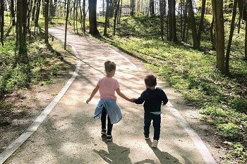 Back view of little girl in pink shirt, blue jacket around waist, holding hands with little boy in dark blue, walking down curving, sunlit trail