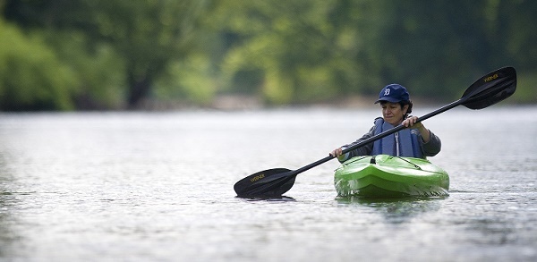 a dark-haired woman in a blue vest and Detroit Tigers cap, paddles a light-green kayak on still, gray-green water. Trees in background