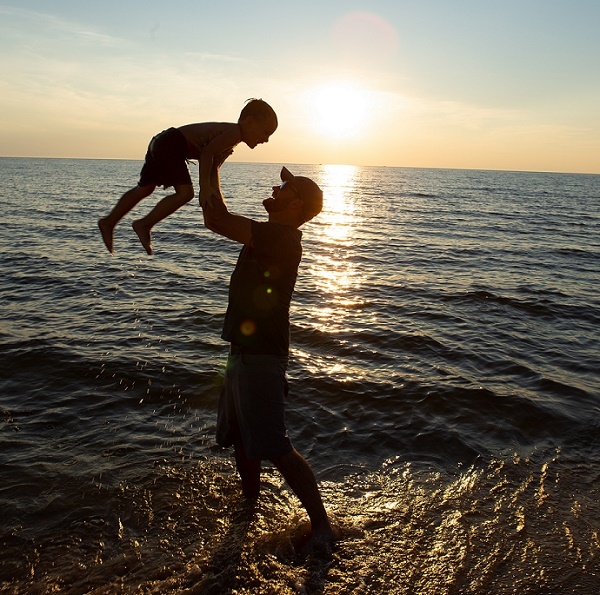 Standing ankle-deep in the shoreline, a man wearing a baseball cap and shorts swings a little boy in the air, backlit by the bright, setting sun. 