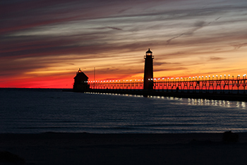 A beautiful orange sunset behind the lighthouse and dock at Grand Haven State Park is shown.