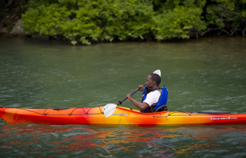 A young person with dark skin wearing a lifejacket operates a red and orange kayak on a sunny day.