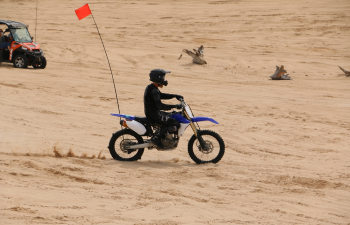 A person with pale skin in dark clothing and a helmet rides a dirt bike across open sand.