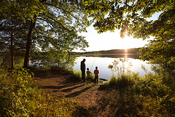 man and two boys fishing on shore of wooded lake at sunset