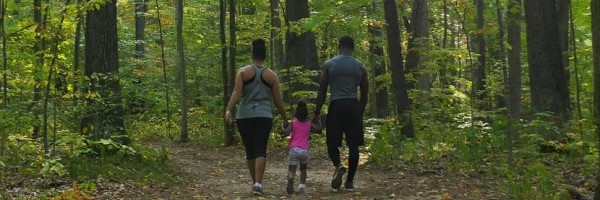 A woman with light brown skin and a man with dark brown walk on either side of a small child with dark skin, each holding one of her hands.