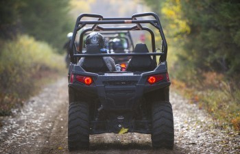 A view of an ORV from the back as it travels down a gravel, forested road. The driver is wearing a silver helmet.