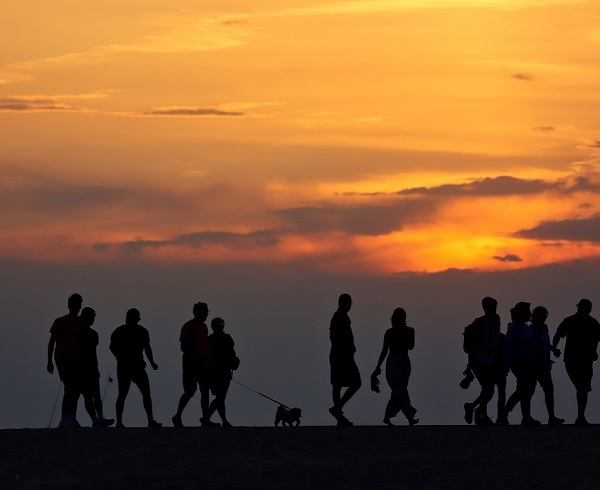 Black shadow outline of about a dozen people walking in line, one walking a dog, along the beach, backlit by a bright, increasingly orange sky