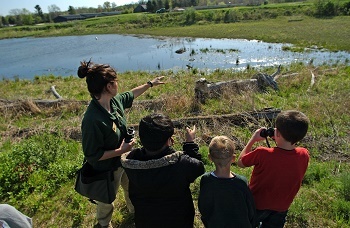 A group of three children stand with a DNR guide, who points across a wetland on a sunny day.