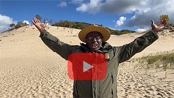 A conservation officer stands in front of a sand dune with arms splayed wide. A red, transparent YouTube play button is overlayed atop the image.