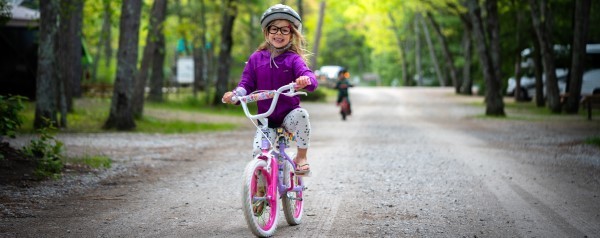 A young child with glasses and long blonde hair rides a bike through a campground, flashing a huge smile to the camera.