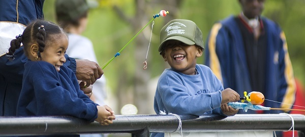 A young Black girl and boy, both laughing and holding fishing poles, lean over the fence on a pier