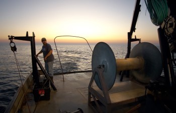 A man stands on the deck of survey vessel Steelhead, silhoutted by the sunset behind him.