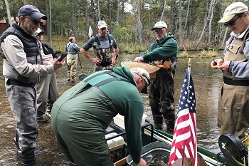 A group of people gather on the Au Sable River with boats, one decorated with an American flag.